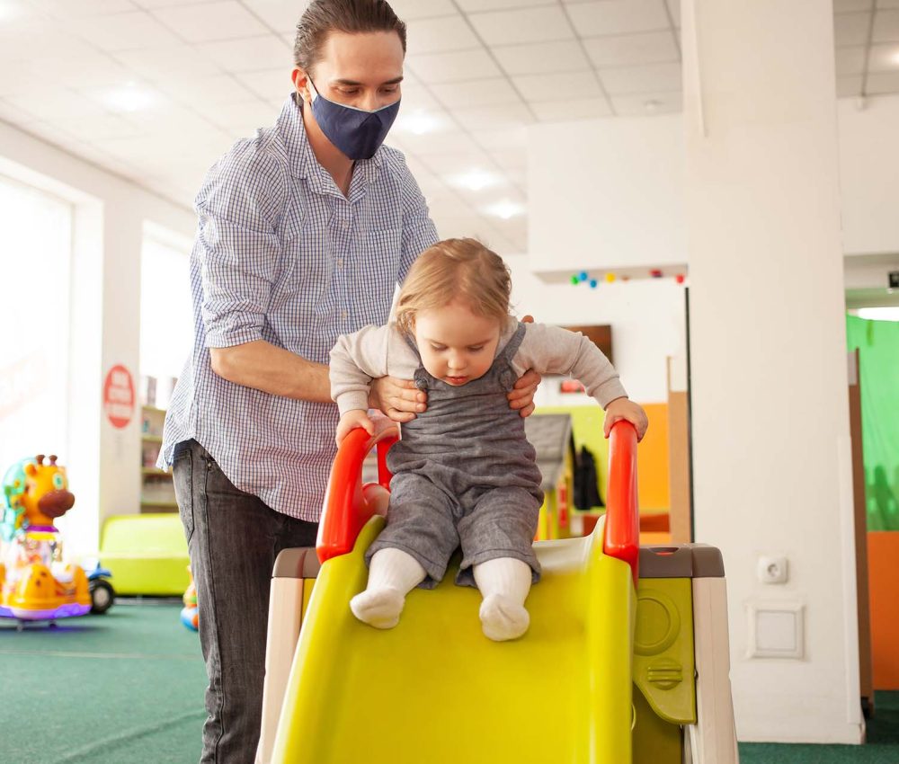 young Dad in a medical mask rolls his daughter from the slide in the game room. Child development center, kindergarten, fun pastime. Family together