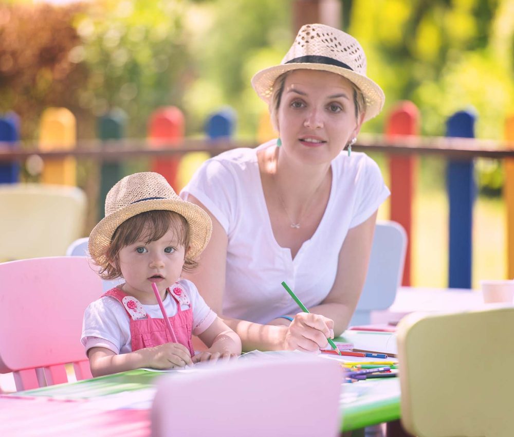 young mother and her little daughter cheerfully spending time together using pencil crayons while drawing a colorful pictures in the outside playschool